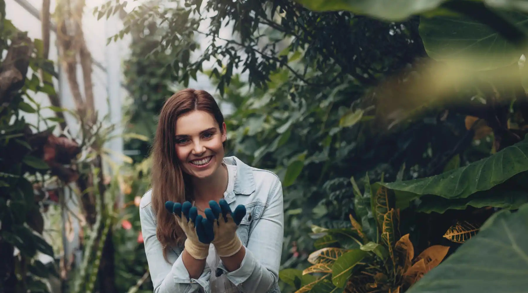 A smiling person in a denim jacket holding something between their hands while surrounded by foliage.